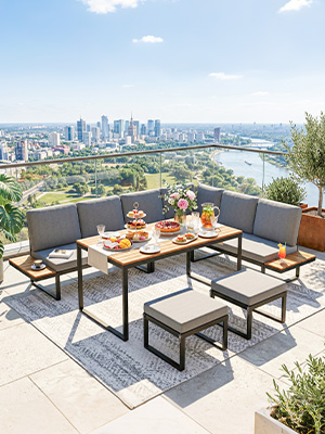 Mobilier d'extérieur moderne installé sur une terrasse surélevée avec paysage urbain et vue sur l'eau en arrière-plan, comprenant un canapé sectionnel à structure en métal noir avec coussins beiges, une table à manger en bois avec couverts et boissons, entourée de plantes en pot.