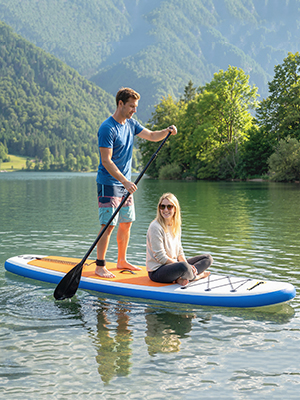 Adulte et enfant sur un paddleboard bleu dans un lac calme entouré de montagnes boisées. L'adulte se tient debout avec une pagaie pendant que l'enfant est assis. Eau claire et paysage pittoresque visibles.