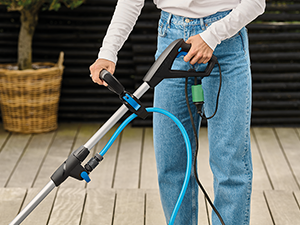 Personne utilisant un aspirateur sans fil sur le plancher d'une terrasse en bois, montrant le bas du corps vêtu d'un jean bleu et l'aspirateur en action.