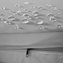 Des gouttelettes d'eau sur une surface grise avec un bord en tissu zippé visible en dessous, suggérant un matériau imperméable ou résistant à l'eau