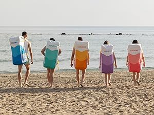 Cinq personnes debout sur la plage face à l'océan, portant des maillots de bain de différentes couleurs en bleu, vert, orange, violet et rose. Vue arrière montrant la disposition coordonnée des couleurs.