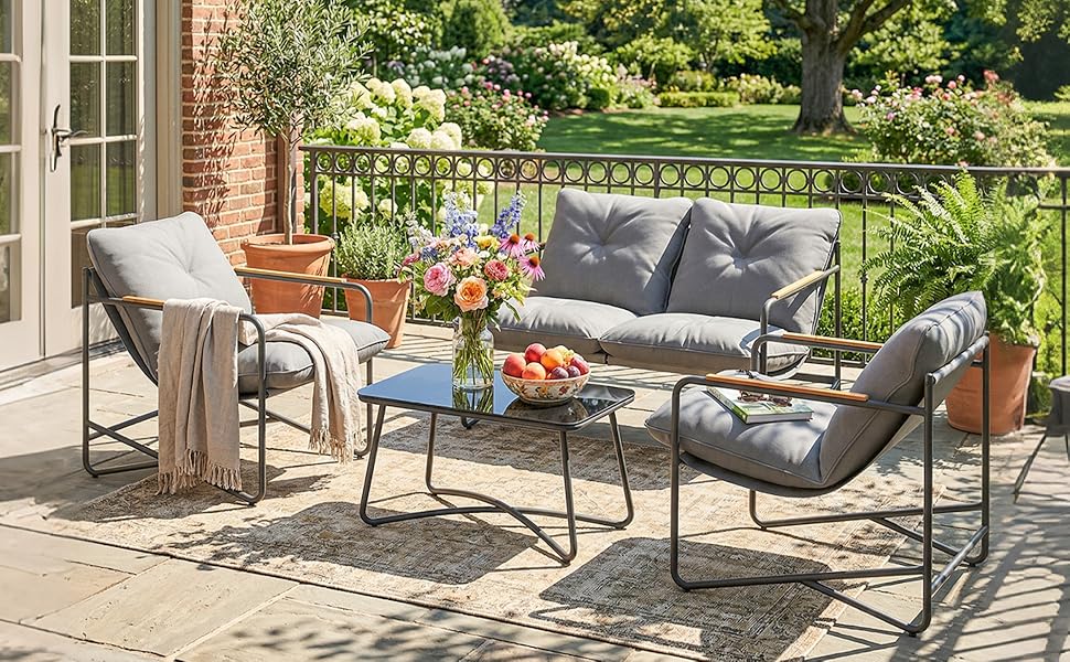 Ensemble de meubles de jardin d'extérieur comprenant un canapé rembourré gris avec des fauteuils assortis, une table basse en verre et une table d'appoint sur un patio en brique avec un jardin luxuriant en toile de fond.