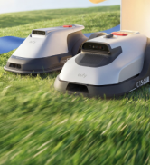 Photographie extérieure montrant une tondeuse à gazon robotisée sur de l'herbe verte avec une station de recharge en arrière-plan. Démonstration d'un équipement d'entretien de jardin autonome dans un environnement résidentiel paysager.