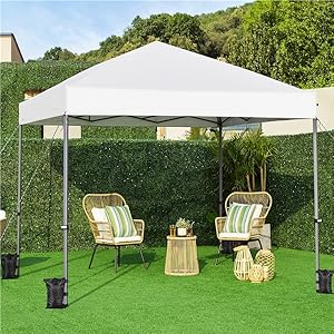Tente à baldaquin escamotable blanche dans un jardin. Deux chaises à rayures vertes et une table en osier en dessous. Entourage de gazon artificiel et de haies.