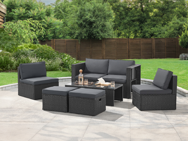 avec des meubles en osier foncé comprenant un canapé, des chaises et un pouf de rangement. Coussins gris. Table avec plateau en verre. Situé sur un patio en pierre avec fond de pelouse verte.