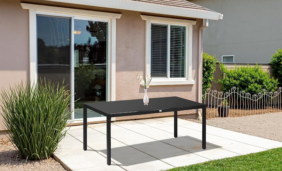 Scène de patio extérieur avec table en métal noir contre mur de maison en stuc beige. Fenêtre avec stores et aménagement paysager désertique visible.