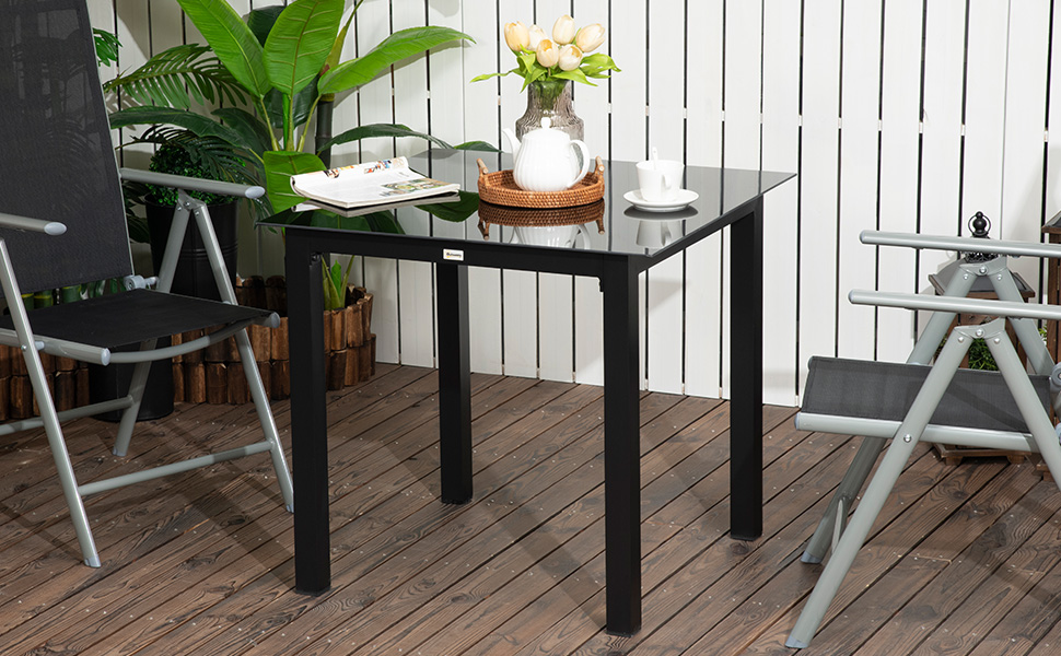 avec plateau en verre et pieds en métal noir, posée sur une terrasse en bois avec chaises pliantes et plantes, adaptée à une utilisation en extérieur