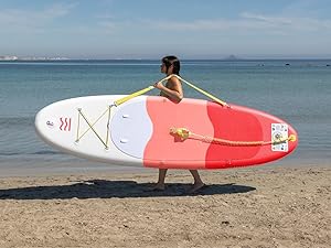 Scène de plage montrant une personne portant un paddleboard de couleur corail au bord de l'eau, avec un océan calme et un horizon en arrière-plan.
