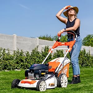 Personne utilisant une tondeuse à gazon orange et noire sur l'herbe dans un jardin extérieur ensoleillé. L'opérateur, vêtu d'un débardeur blanc et d'un chapeau brun, pousse activement une tondeuse automotrice sur la pelouse.