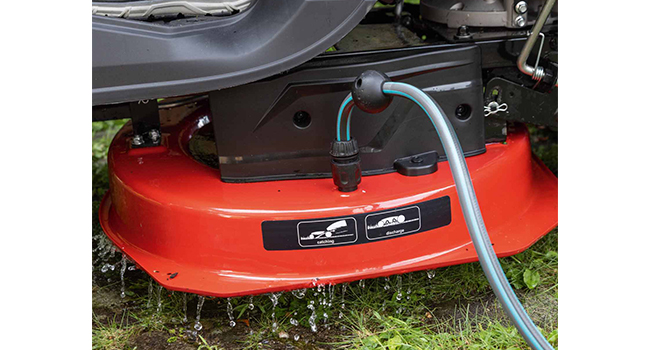 Unité de base de tondeuse à gazon robotisée rouge avec connexion de charge visible, positionnée sur l'herbe avec les composants mécaniques visibles.