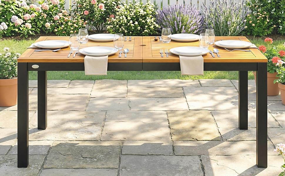 Table à manger extérieure avec structure en métal noir et surface supérieure en bois, positionnée sur un trottoir en béton avec aménagement paysager visible.